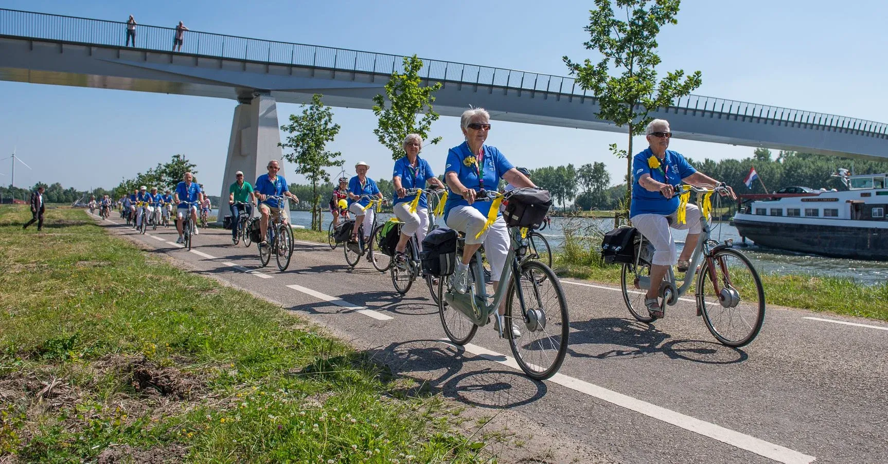 Een lange rij recreanten in blauwe poloshirts met gele linten fietst over een fietspad langs het kanaal, onder een fietsbrug door
