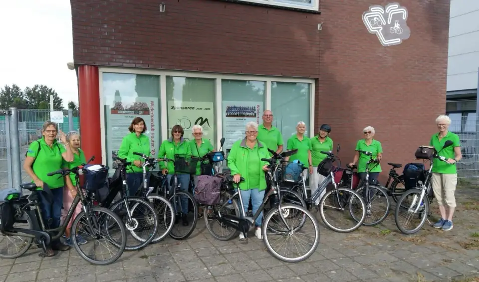 Recreanten in groene poloshirts en jassen met hun fietsen voor het clubhuis, met het clublogo op de gevel