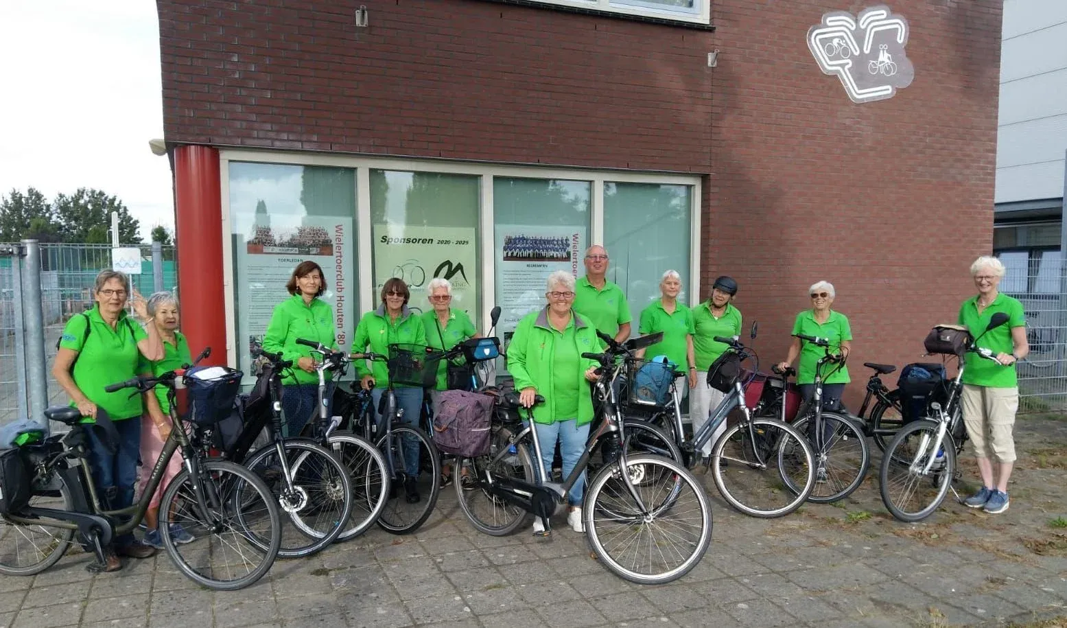 Recreanten in groene poloshirts en jassen met hun fietsen voor het clubhuis, met het clublogo op de gevel