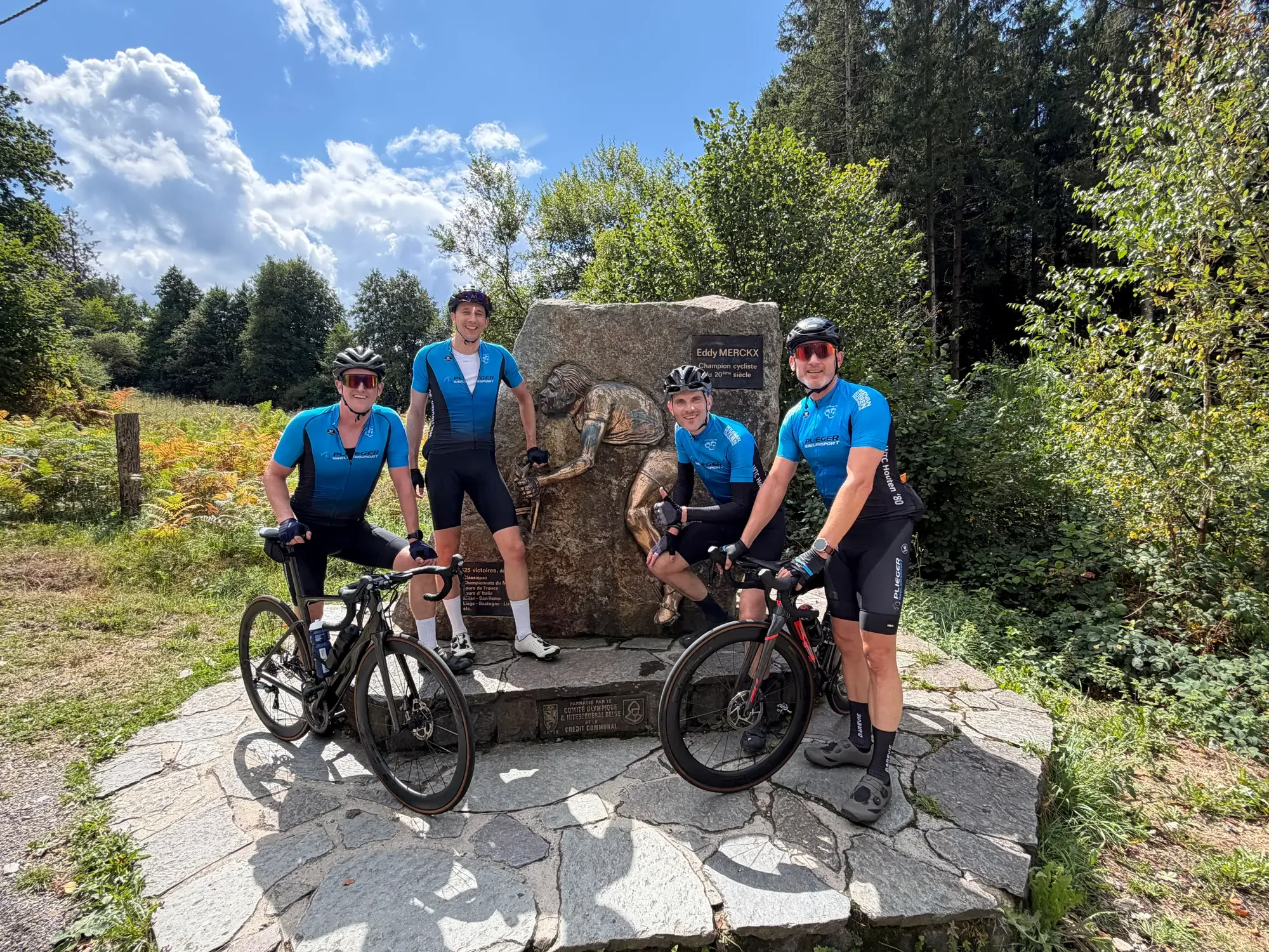 Vier toerleden in het blauwe clubtenue met hun racefietsen bij een wielermonument in de Ardennen