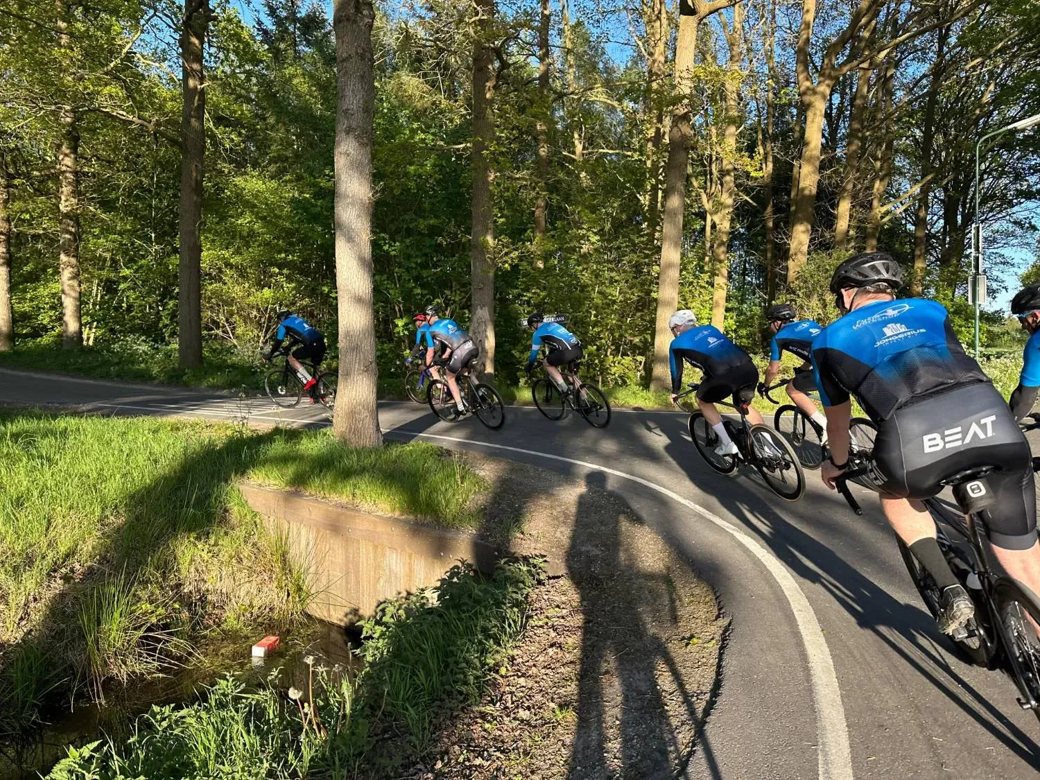 Toerleden in het blauwe clubtenue fietsen achter elkaar door een bocht langs de bosrand, in de avondzon