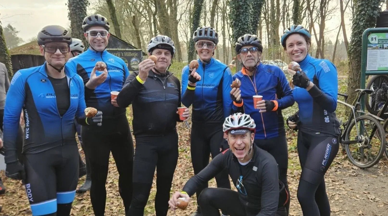 Lachende toerleden in winterkleding en met helm houden tijdens een gravelrit in het bos een oliebol omhoog