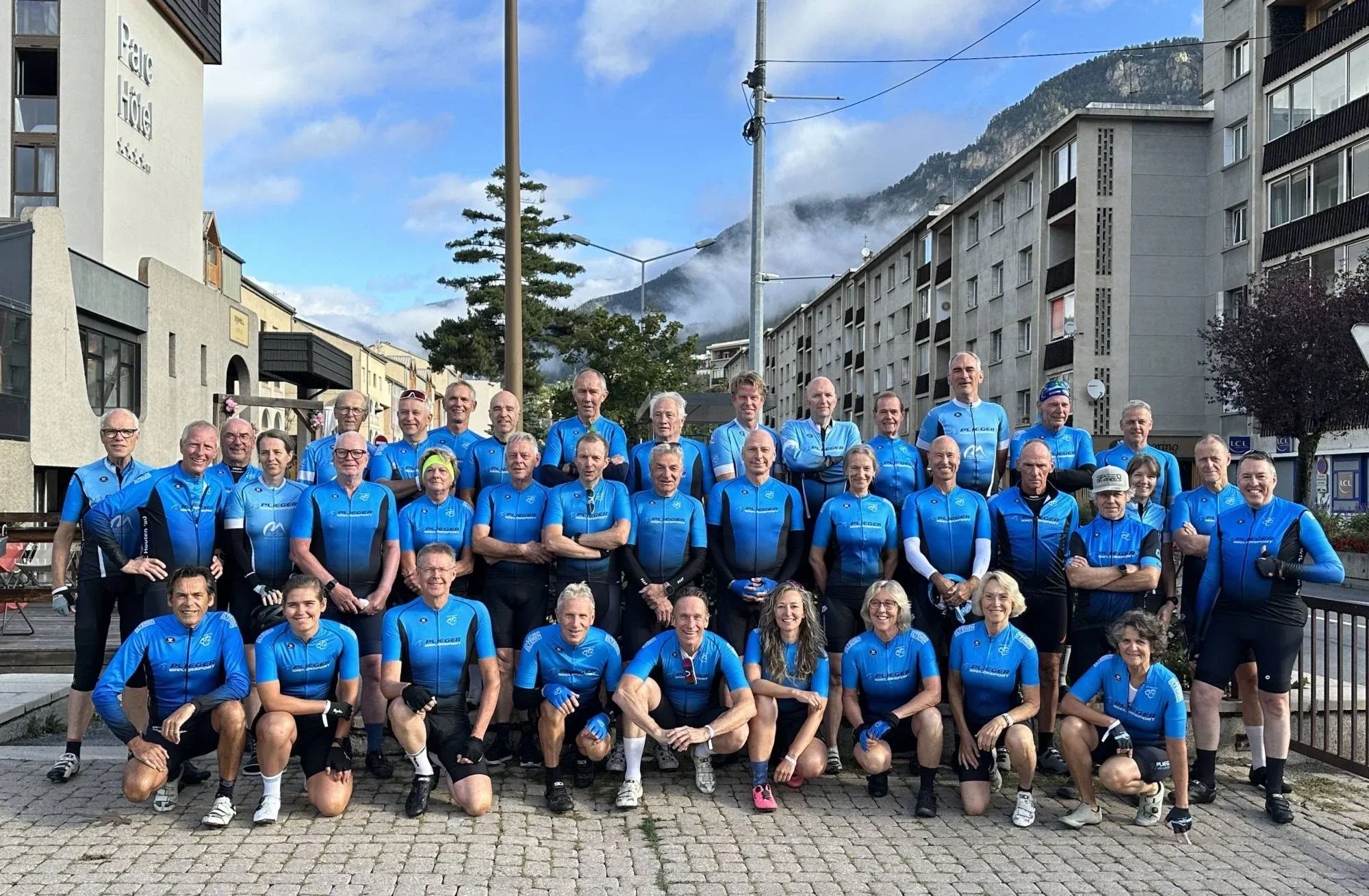 Groepsfoto van ruim veertig toerleden in het blauwe clubtenue in een straat, met beboste bergen en laaghangende wolken op de achtergrond