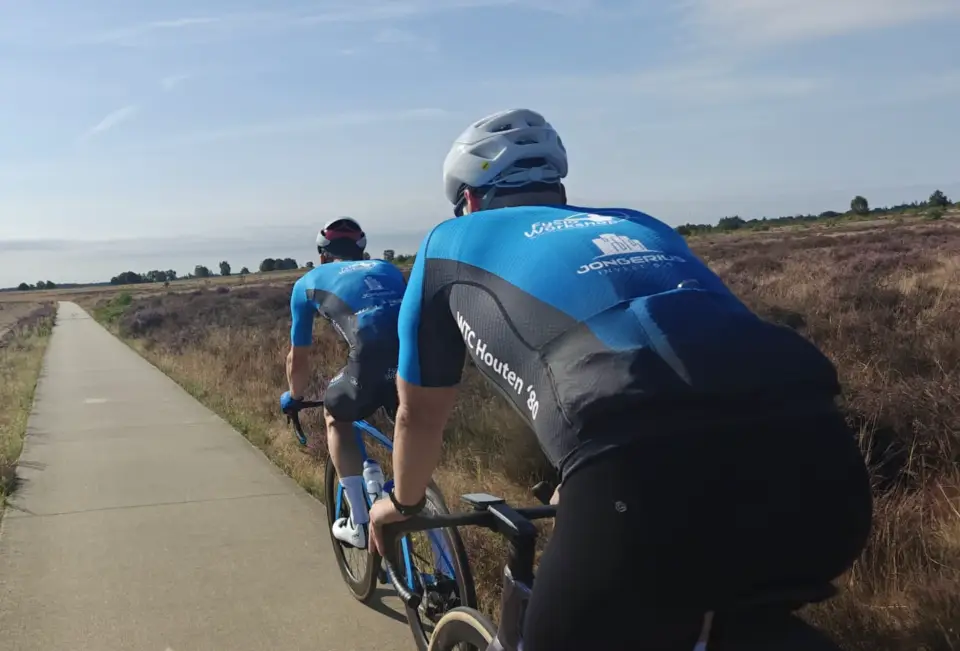 Twee toerleden van achteren gezien in het blauwe clubtenue, achter elkaar op een fietspad door de bloeiende heide