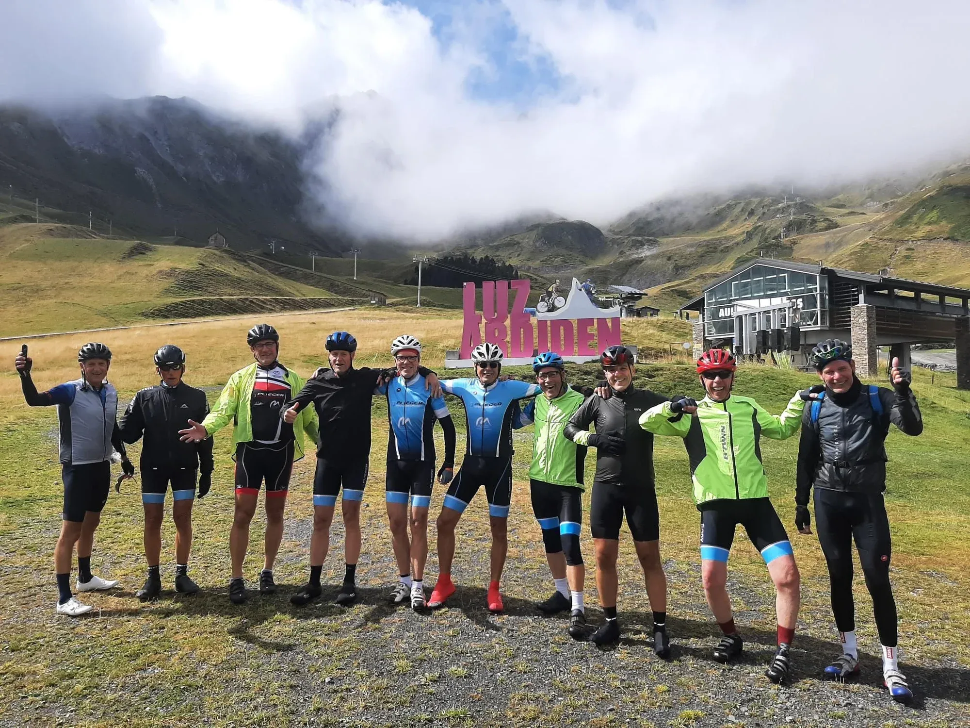 Een groep toerleden arm in arm bij het bord van Luz Ardiden in de Pyreneeën, met wolken boven de bergen