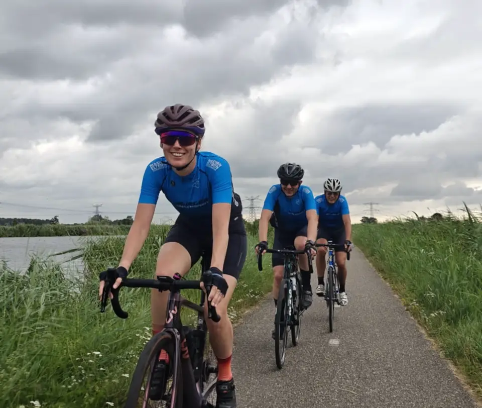 Drie lachende toerleden in het blauwe clubtenue fietsen achter elkaar over een smal fietspad langs het water, onder een bewolkte lucht