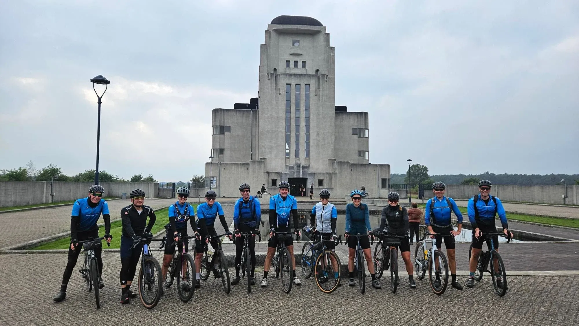 Twaalf fietsers, de meesten in het blauwe clubtenue, op een rij met hun gravelfietsen voor het gebouw van Radio Kootwijk