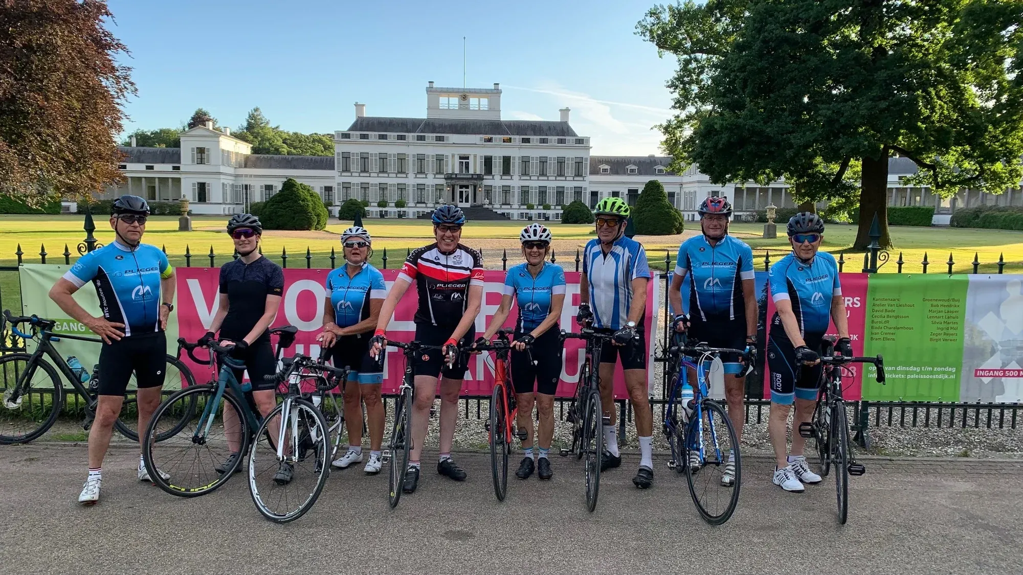 Acht toerleden met helm staan naast elkaar met hun racefietsen voor Paleis Soestdijk