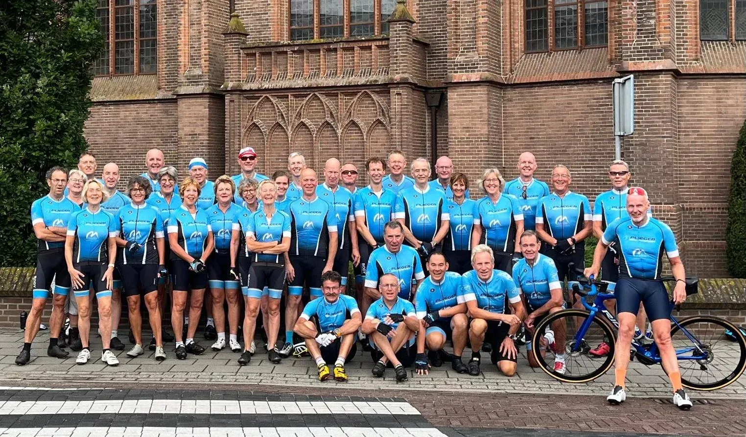 Groepsfoto van ruim veertig lachende toerleden in het blauwe clubtenue voor een bakstenen kerk