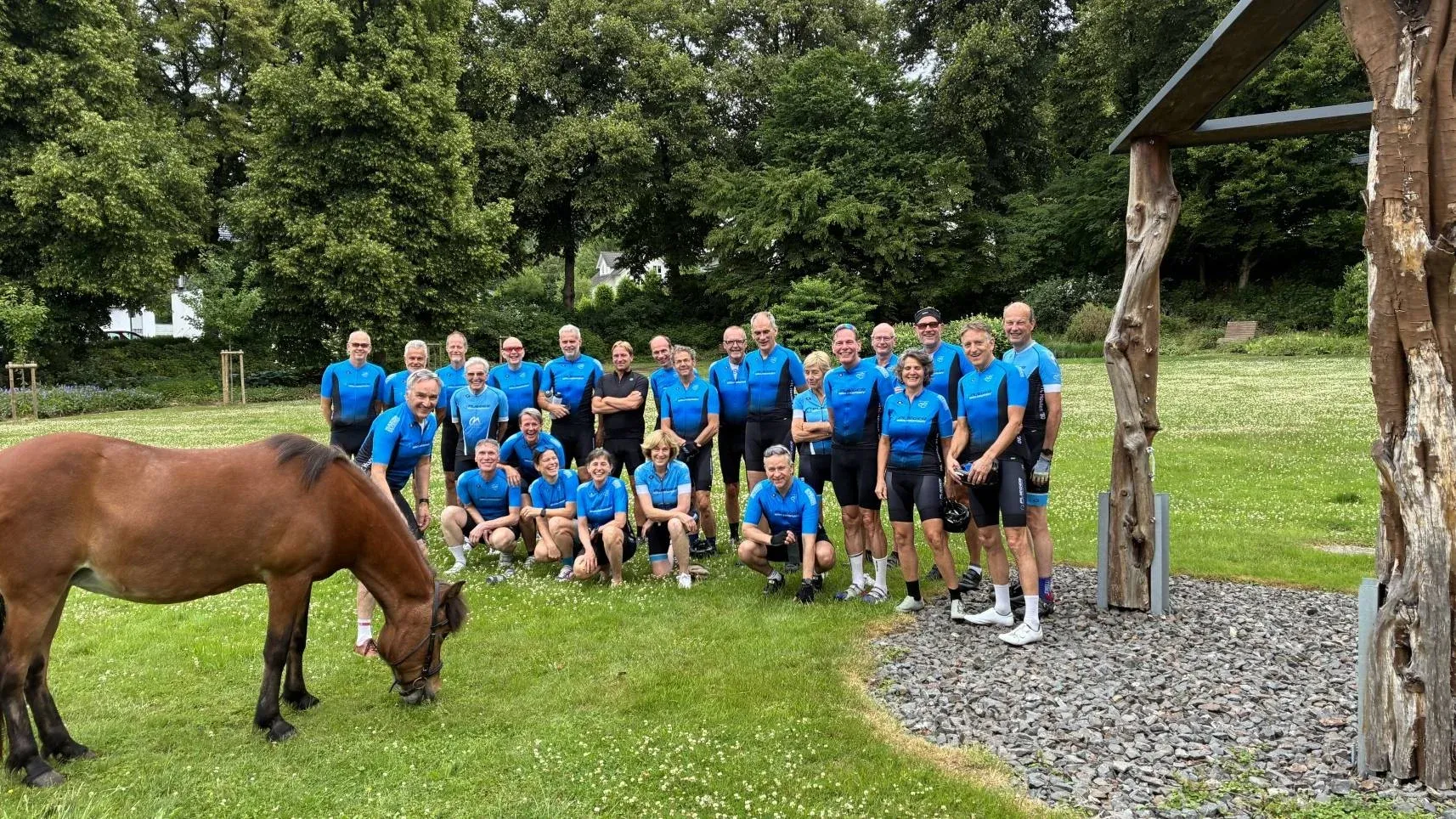 Groepsfoto van toerleden in het blauwe clubtenue op een grasveld, met op de voorgrond een grazend paard