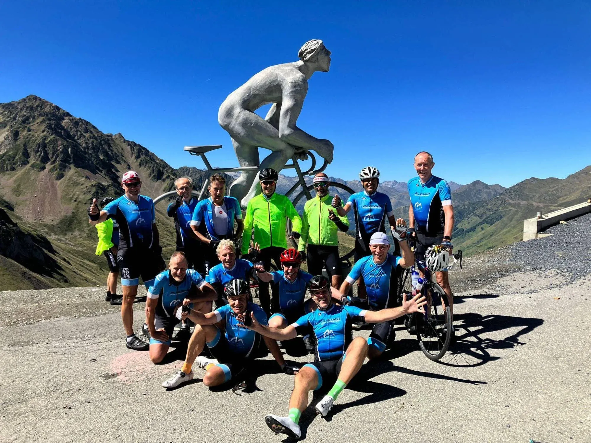 Juichende toerleden in blauwe clubshirts bij het fietsersbeeld op de top van de Col du Tourmalet, met de bergtoppen van de Pyreneeën erachter