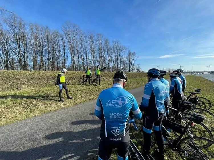Toerleden in het clubtenue staan met hun racefietsen op een dijk en krijgen uitleg van drie fietsers in gele hesjes
