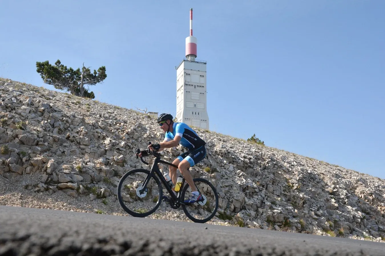Een toerlid in het clubtenue klimt op de racefiets langs de kale stenen helling van de Mont Ventoux, met de zendmast op de top erachter